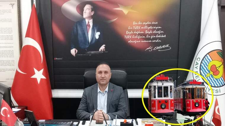 Masasında oturan bir kişi, Türk bayrağı ve itfaiye araçlarının yer aldığı bir pano önünde görülüyor.