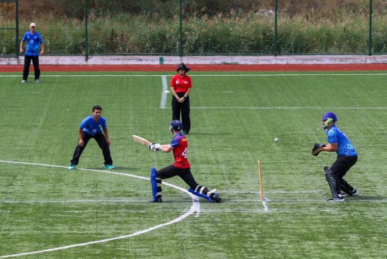 Suni çim sahada kriket oynayan bir sporcu, sopasını uzatırken arkasında başka bir oyuncu çömeliyor.