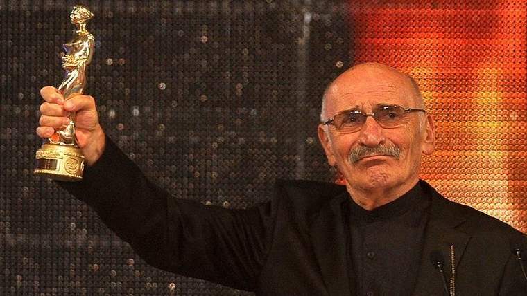 Bir adam, elinde ödül tutarak gülümsüyor. Arkada ışıklar parlıyor.
