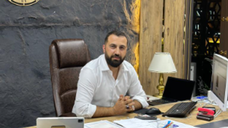 Bir masa başında oturan bir kişi, önünde bilgisayar ve belgeler var.