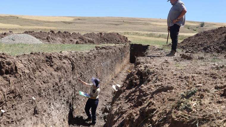 Bir kişi, toprakta açılmış bir hendekte ölçüm yaparken, diğer kişi yukarıda duruyor.