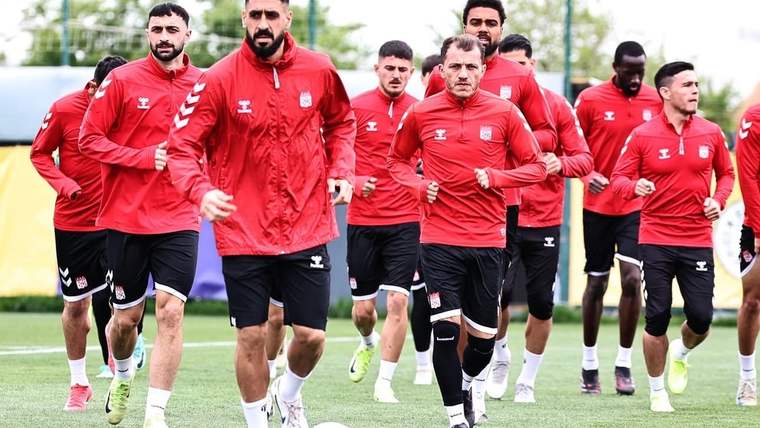 Kırmızı antrenman kıyafetleri giyen futbolcular, çim sahada yan yana koşarken görülüyor.
