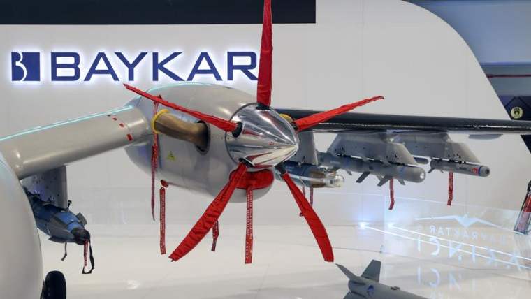 Baykar logosunun arkasında, kırmızı iplerle kaplı bir motor ve yanındaki silahlar sergileniyor.