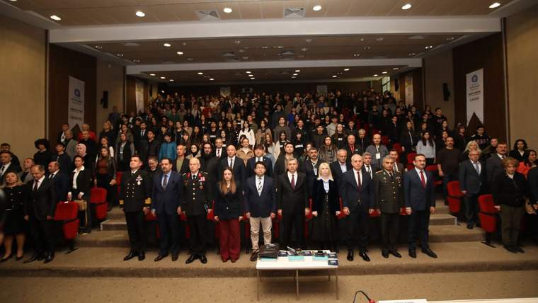 Konferans salonunda, ön sırada duran kalabalık bir grup ile arkalarında oturan izleyiciler görülüyor.