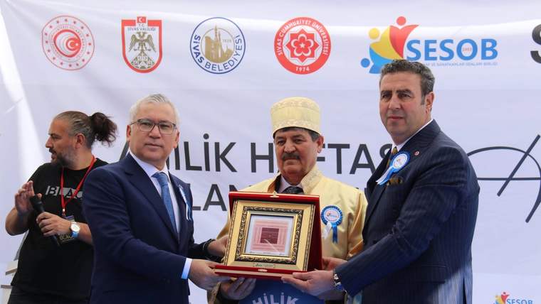 Üç kişi, ödül takdiminde bulunuyor; biri ödülü tutarken diğerleri gülümsüyor.