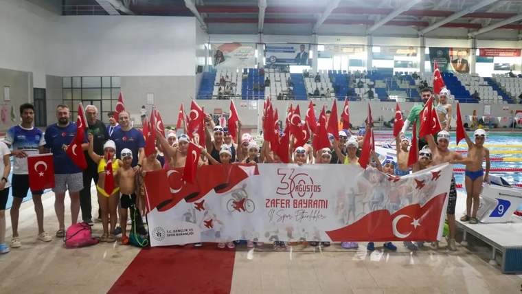 Kapalı yüzme havuzunda çocuk ve yetişkinler, Türk bayrakları ve 30 Ağustos Zafer Bayramı pankartıyla poz veriyor.