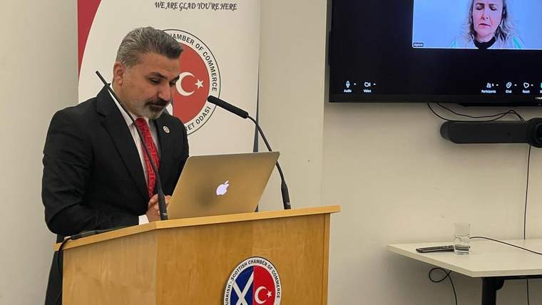 Kürsüde dizüstü bilgisayarla konuşan bir kişi ve ekranda görüntülü görüşmeye katılan bir kadın görülüyor.