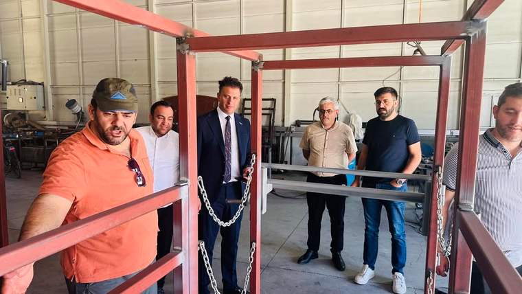 Atölyede kırmızı metal bir konstrüksiyonun çevresinde duran ve inceleyen beş erkek görülüyor.