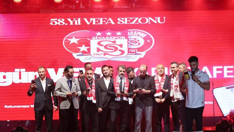 Kırmızı sahnede Sivasspor logoları önünde atkılı ve takım elbiseli bir grup insan yan yana duruyor.