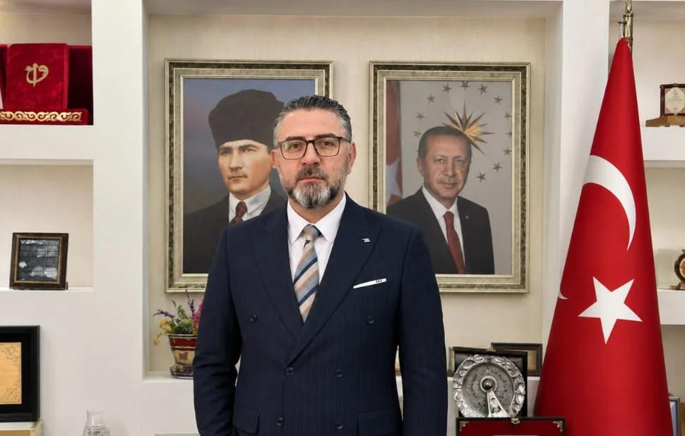 Takım elbiseli sakallı bir adam, arkasında iki portre ve Türk bayrağı bulunan bir odada duruyor.