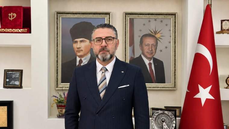 Takım elbiseli sakallı bir adam, arkasında iki portre ve Türk bayrağı bulunan bir odada duruyor.