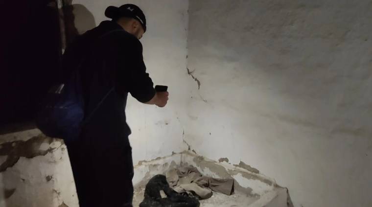 Karanlık ve harap bir odada, el feneri tutan siyah giysili bir kişi çatlamış duvarların önünde duruyor.