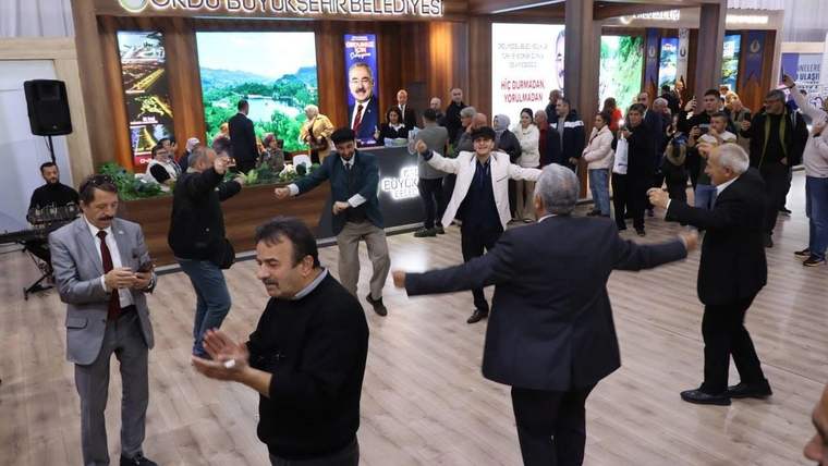 Bir grup insan, bir etkinlikte müzik eşliğinde dans ediyor. Arka planda tanıtım stantları yer alıyor.