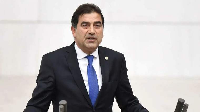 Koyu takım elbiseli bir kişi, kürsü önünde mikrofona benzer cihazların arasında konuşuyor.