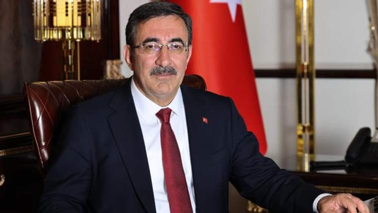 Kırmızı kravat takmış bir adam, deri koltukta oturuyor ve arkasında Türk bayrağı var.