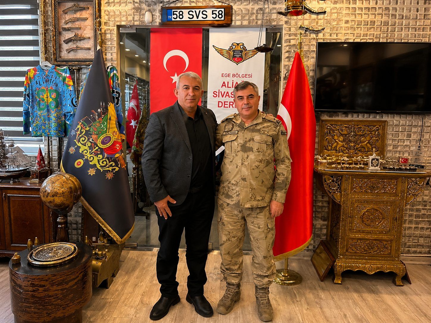İç mekânda iki erkek, Türk bayrağı ve jandarma flaması önünde yan yana duruyor; çevrede süslü eşyalar bulunuyor.