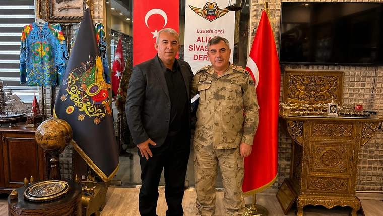 İç mekânda iki erkek, Türk bayrağı ve jandarma flaması önünde yan yana duruyor; çevrede süslü eşyalar bulunuyor.