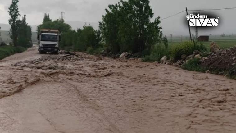 Çamurlu sel sularıyla kaplı yolda bir kamyon, ağaçlar ve tarlaların arasında ilerliyor.