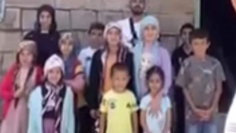 Bir grup çocuk, gülümseyerek kameraya poz veriyor. Arka planda taş duvar görünmekte.
