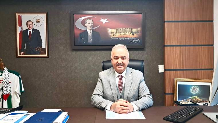 Takım elbiseli bir kişi, arkasında portreler ve bayrak bulunan odada masasının başında oturuyor.