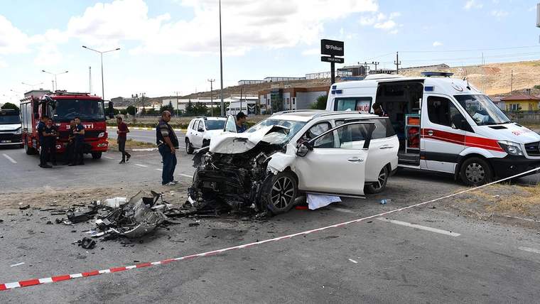 Hasar görmüş beyaz otomobil, itfaiye ve ambulans araçlarının bulunduğu yolda güvenlik şeridiyle çevrilmiş.