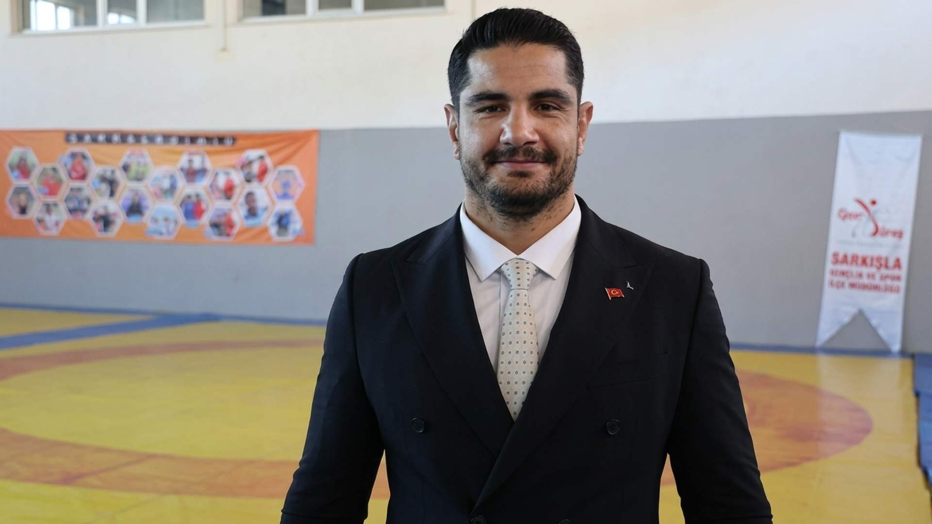 Spor salonunda takım elbiseli, kravatlı ve rozetli bir erkek kameraya bakarak ayakta duruyor.