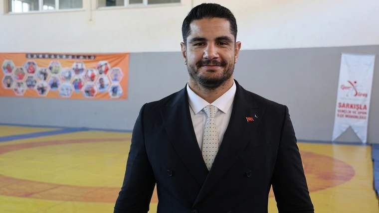 Spor salonunda takım elbiseli, kravatlı ve rozetli bir erkek kameraya bakarak ayakta duruyor.