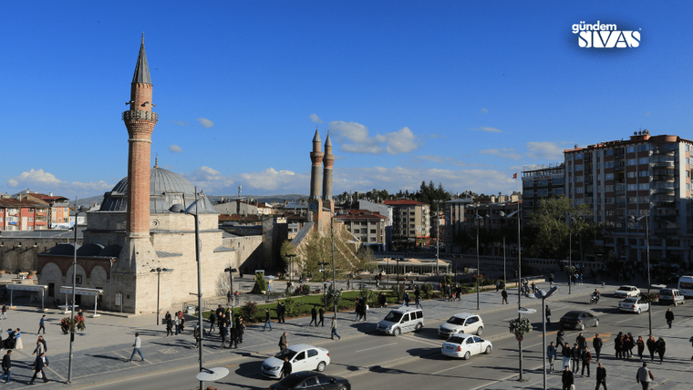Minareli tarihi bir cami, çevresindeki kent meydanı, araçlar ve yayalarla birlikte görülüyor.