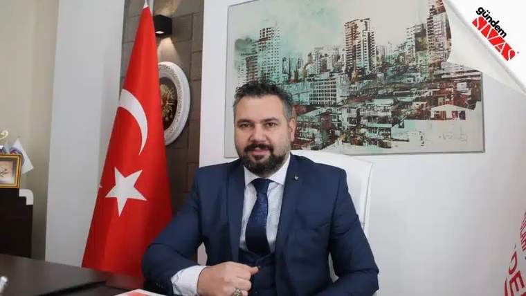 Masa başında oturan takım elbiseli bir kişi, yanında Türk bayrağı ve şehir silüeti bulunan tabloyla görülüyor.