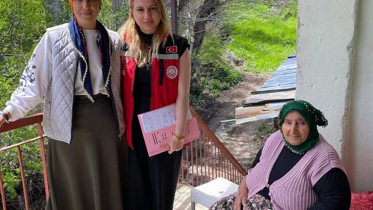Balkonda iki kadın ayakta dururken, yeşil başörtülü yaşlı bir kadın sandalyede oturuyor.