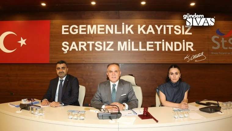 Bir toplantı odasında, masada oturan üç kişi, arka planda Türk bayrağı ve yazı ile birlikte görünüyor.