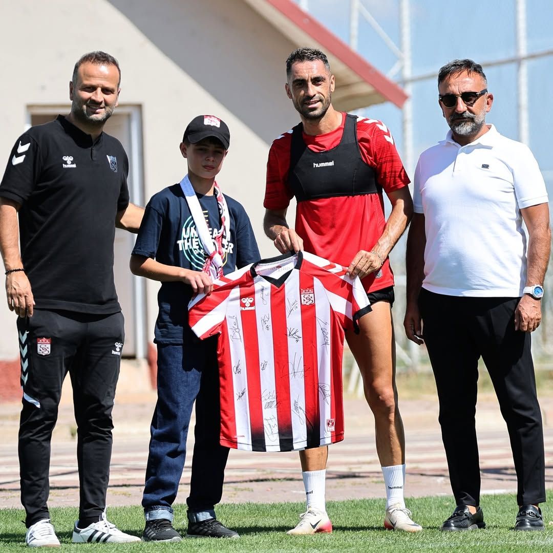 Üç yetişkin ve bir çocuğun önünde, kırmızı-beyaz çizgili futbol formasını tutan iki kişi görülüyor.