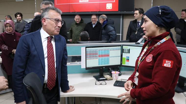 Bir ofiste, bilgisayar ekranlarının önünde bir kişiyle konuşan bir kadın, arka planda kalabalık bir grup var.
