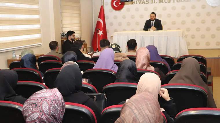 Bir toplantı salonunda, masanın önünde bir kişi konuşuyor; dinleyiciler arasında kadınlar ve erkekler var.