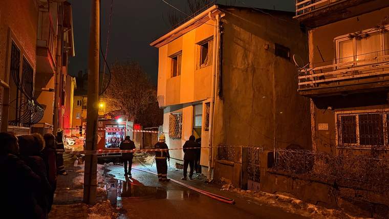 Gece, iki katlı bir evin önünde acil durum aracı, görevliler ve çevrede bekleyen insanlar görülüyor.