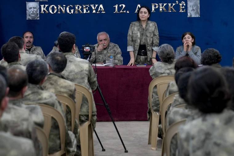 Kamuflaj giysili kişilerin bulunduğu salonda bir kadın masanın önünde konuşurken görülüyor.