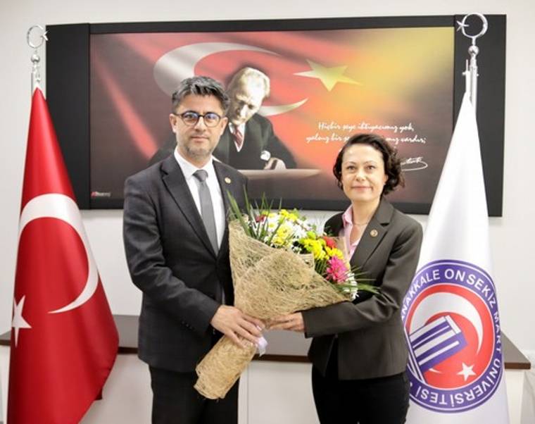 Bir erkek ve bir kadın, Türk bayrağı ile Çanakkale Onsekiz Mart Üniversitesi bayrağı önünde çiçek tutuyor.