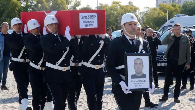 Beyaz miğferli üniformalı görevliler, üzerinde Cuma Şimşek yazan kırmızı tabutu taşırken ilerliyor.