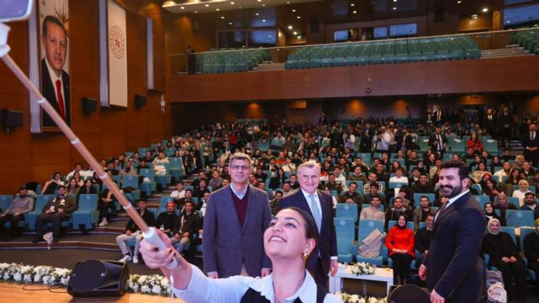 Bir kadın, selfie çubuğu ile fotoğraf çekiyor; arka planda kalabalık bir izleyici ve sahnede üç kişi var.
