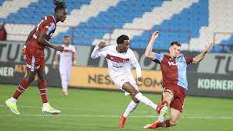 Yeşil sahada beyaz formalı futbolcu topa vururken iki kırmızı formalı oyuncu müdahale ediyor.