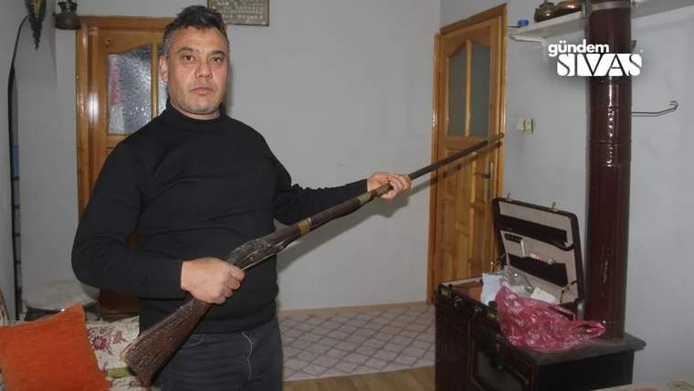 Bir adam, elinde uzun bir tüfek tutuyor; arka planda bir kapı ve ocak görünüyor.