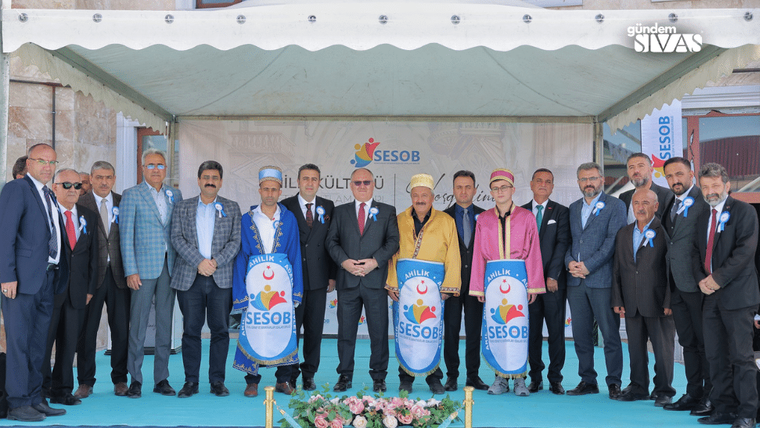 Sahne önünde, geleneksel kıyafetler giymiş bir grup erkek ve kadın, yan yana duruyor.