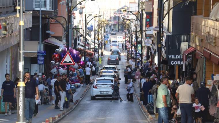 Dükkânlarla çevrili caddede araçlar ilerlerken kaldırımlarda yayalar ve renkli balonlar görülüyor.