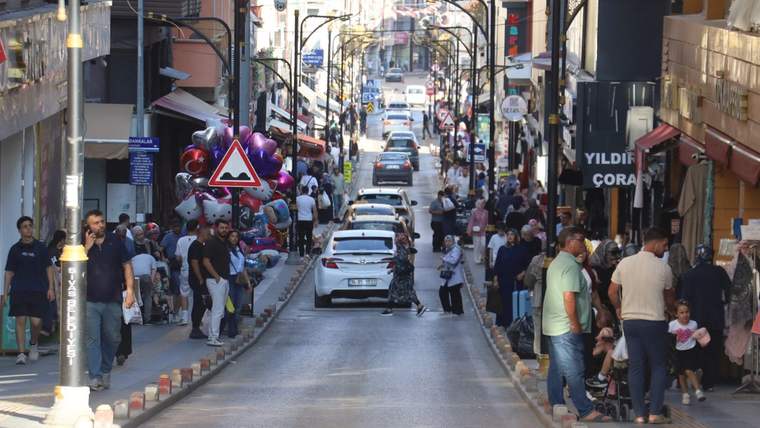 Dükkânlarla çevrili caddede araçlar ilerlerken kaldırımlarda yayalar ve renkli balonlar görülüyor.