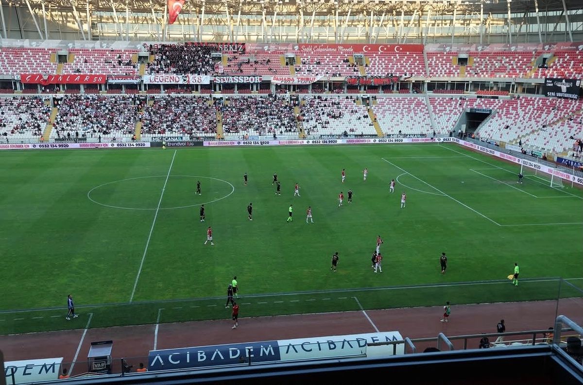 Futbol sahasında oyuncular mücadele ederken tribünlerin büyük bölümü boş görünüyor.