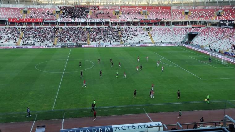 Futbol sahasında oyuncular mücadele ederken tribünlerin büyük bölümü boş görünüyor.