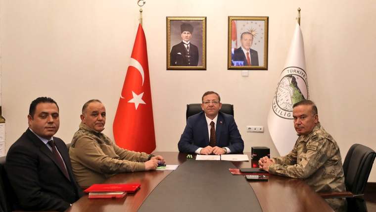 Masa etrafında oturan dört kişi, arkalarında Türk bayrağı, iki portre ve duvar amblemiyle görülüyor.