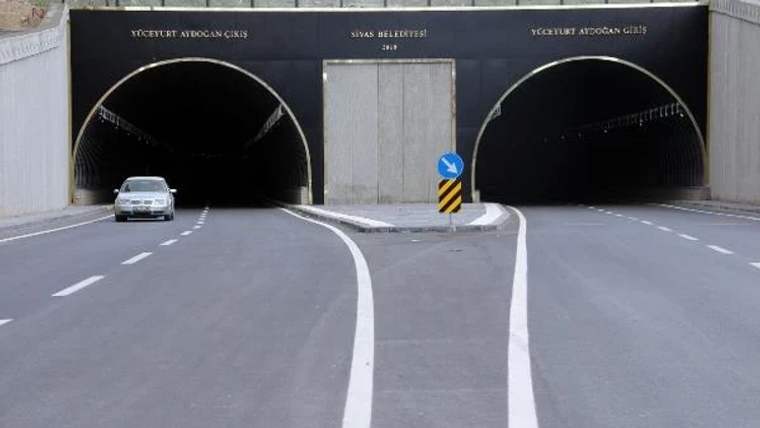 İki tünel girişinin önünde bölünmüş yol, yön levhası ve tek bir otomobil görülüyor.