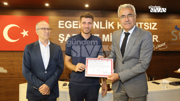 Bir kişi, elinde sertifika tutarak başka bir kişiyle tokalaşıyor; arka planda Türk bayrağı ve yazılı pano var.