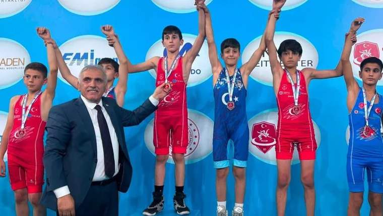 Üzerinde madalya bulunan iki güreşçi, ellerini havaya kaldırmış; yanlarında bir yetişkin kişi var.